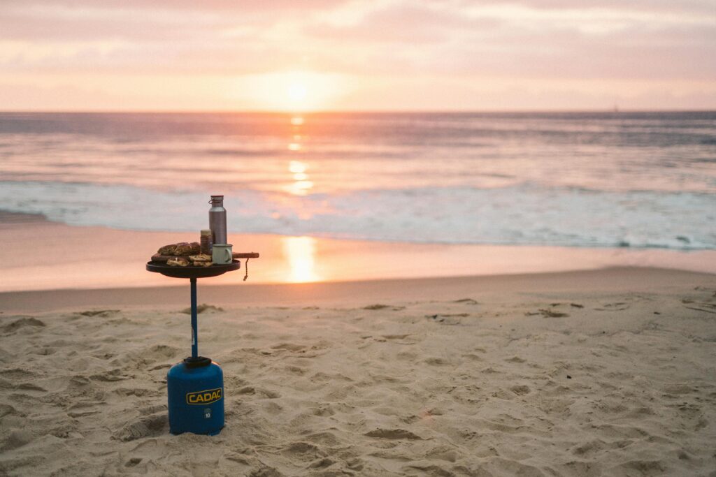 pexels-photo-9716779-9716779 A portable camping stove on a sandy beach at sunrise with food and drink, capturing an adventurous outdoor vibe.